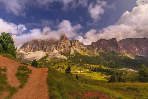 Camino de tierra rojiza que se adentra en un valle alpino con imponentes montañas cubiertas de nubes. La ruta Cinque Torri se revela entre prados verdes y bosques frondosos.