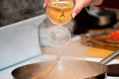 Eine Hand gießt Weißwein aus einem Glas in eine dampfende Pfanne. Das Verkaufen von Wein beim Kochen verleiht dem Gericht Tiefe.