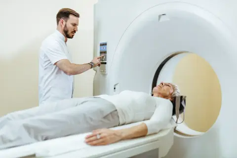 Pacjentka leży na stole tomografu, technik przygotowuje badanie. Czy można pić wodę przed tomografią z kontrastem? Zapytaj lekarza.