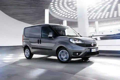 Szary Fiat Doblo Cargo w ruchu, z rozmytymi kołami, na tle nowoczesnej architektury z okrągłymi światłami.