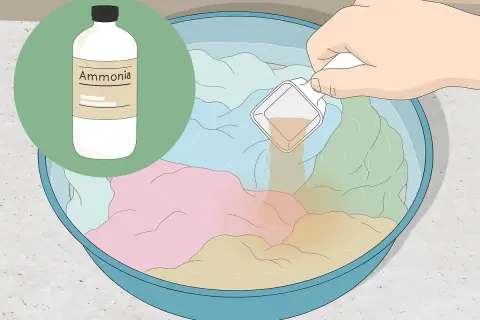Eine Hand gibt eine Messlöffel-Menge einer Flüssigkeit in eine Schüssel mit bunten Kleidungsstücken und Wasser. Die Flüssigkeit hilft, den **Geruch aus Kleidung entfernen**.