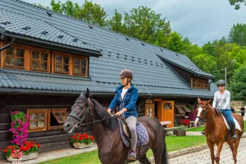Ludzie jeżdżą konno przed drewnianym budynkiem stadniny.