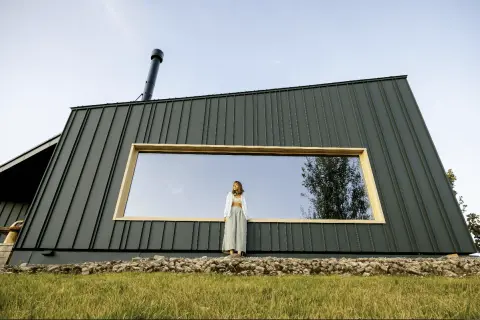 Kobieta stoi przed nowoczesnym domem pokrytym ciemną blachą na rąbek. Duże okno odbija niebo i drzewa.