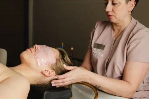 Relaks w gabinecie head spa. Kobieta leży z kamieniami na oczach, a masażystka delikatnie dotyka jej skroni.