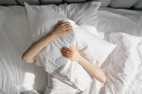 A person clutches a pillow in bed, sunlight dappling the sheets. This scene evokes the struggle many with ADHD face: do people with ADHD have trouble sleeping?