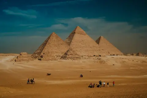 Pustynia Giza z piramidą Cheopsa w tle. Ludzie i wielbłądy przemierzają piaski pod błękitnym niebem.