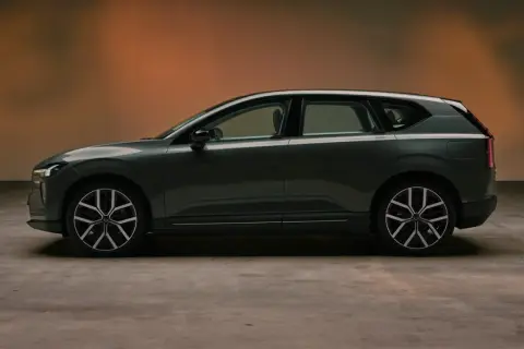 Volvo EX60 gris oscuro, perfil lateral, con fondo degradado naranja y suelo de hormigón.