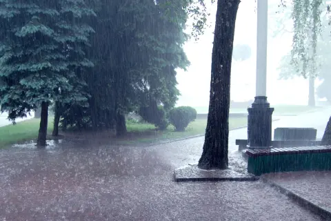 Intensywny deszcz nawalny zalewa parkową alejkę i trawnik. Drzewa pochylają się pod naporem wody.