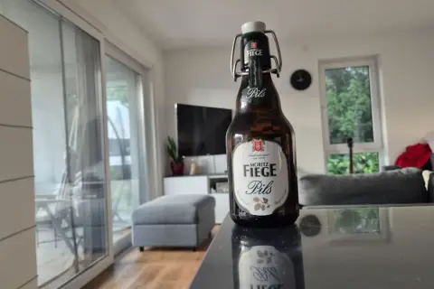 Eine Flasche Moritz Fiege Pils steht auf einem Tisch. Im Hintergrund ein Wohnzimmer mit Sofa und Fernseher.