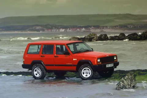 Czerwony Jeep ZJ na kamienistej plaży, z widokiem na morze i wzgórza w tle.