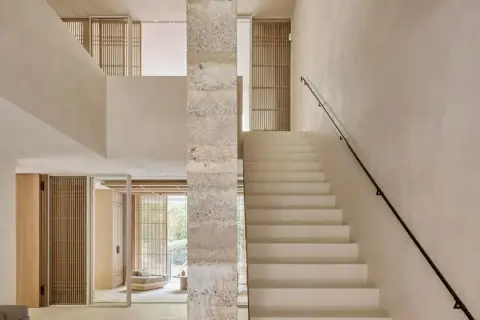 Escaleras de cemento blancas y minimalistas ascienden junto a un pilar de piedra texturizada, conectando niveles en un interior luminoso.
