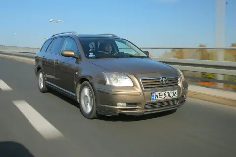 Brązowy Toyota Avensis T25 w kombi jedzie po drodze. Widać przednie żarówki i tablicę rejestracyjną WE 80036.