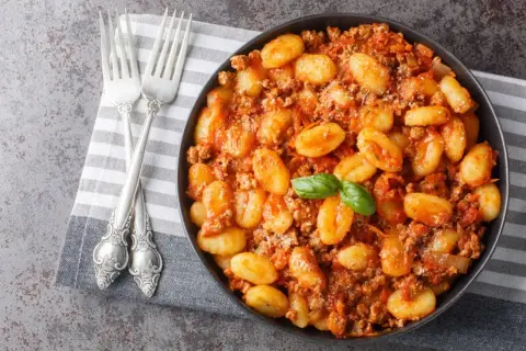 Un plat de gnocchi maison savoureux, nappés d'une sauce bolognaise riche et garnis d'une feuille de basilic frais. Deux fourchettes argentées sont posées sur une serviette à côté.