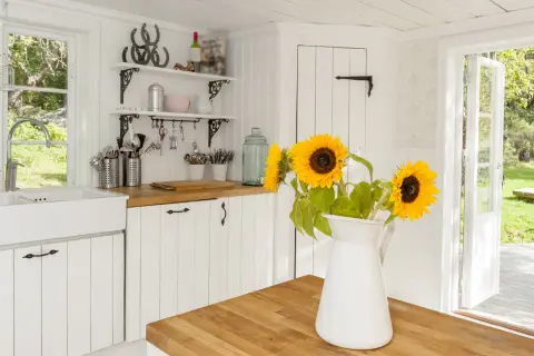 Helle Küche im modernen Landhausstil mit Spülbecken, Holzarbeitsplatte und Sonnenblumen. Offene Tür gibt Blick auf grünen Garten frei.