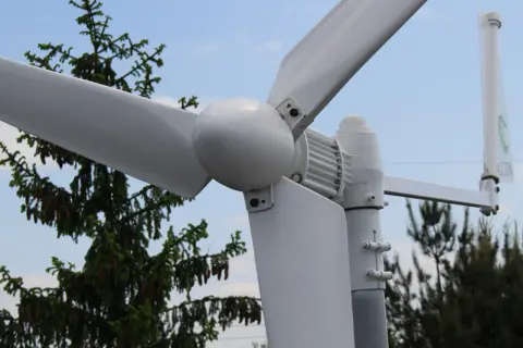 Montaż elektrowni wiatrowej w pobliżu drzew. Białe łopaty wirnika i gondola na tle błękitnego nieba.