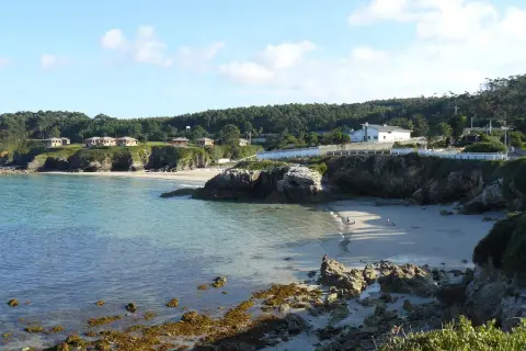 La **playa Burela** se asoma entre rocas y acantilados, con casitas blancas coronando la colina. El mar turquesa acaricia la arena dorada donde pasean algunas personas.