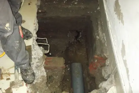 Excavación en el suelo de una vivienda para acceder a una arqueta con tuberías. Se ven restos de ladrillo y tierra.