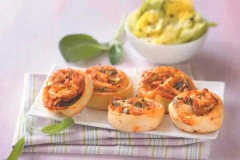 Fünf leckere Pizzaschnecken Thunfisch mit Tomaten und Lauchzwiebeln auf einem weißen Teller, daneben ein Schälchen mit gelbem Dip.