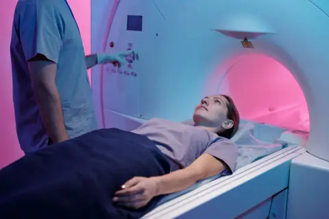 Une femme est allongée dans un appareil de TDM. Le technicien, en blouse bleue, ajuste les commandes. La TDM, c'est quoi ? Un examen d'imagerie médicale.