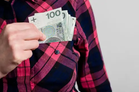 Ręka wkłada banknoty 100 zł do kieszeni koszuli w kratę. Może to symbolizować wynagrodzenie ryczałtowe.