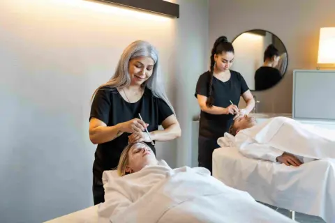Dos terapeutas aplican tratamientos faciales a dos clientes en camillas. Una mujer con cabello plateado realiza un **olimpia masaje** facial a una cliente rubia.