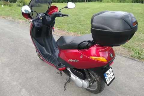 Czerwony skuter Peugeot Elyseo 125 z czarnym kufrem na bagaż, gotowy na podróż po malowniczych drogach.