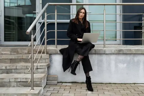 Elegancka kobieta w czarnym płaszczu, z laptopem na kolanach, prezentuje stylowe dodatki: wzorzyste rajstopy i sznurowane botki.