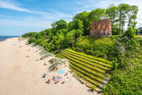 Piękny klif w Trzęsaczu z ruinami kościoła na szczycie. Piaszczysta plaża z wypoczywającymi ludźmi poniżej.