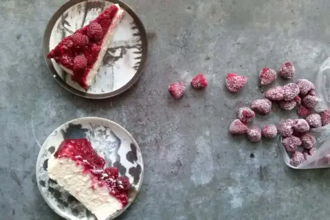 Dwa kawałki kremowego sernika z malinami na talerzach, obok garść mrożonych malin. Idealny deser na letnie popołudnie.