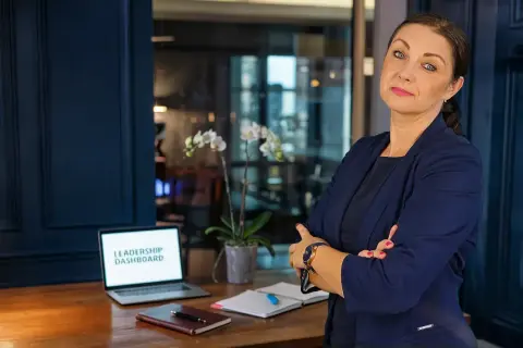 Kobieta w garniturze, z laptopem z napisem "LEADERSHIP DASHBOARD", uczy jak zostać managerem.