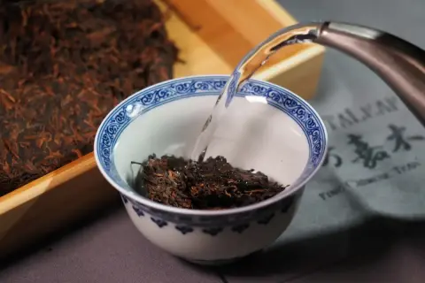 Préparation d'un thé de Chine. De l'eau chaude est versée dans un bol rempli de feuilles de thé sombres, à côté d'un bloc de thé compressé.
