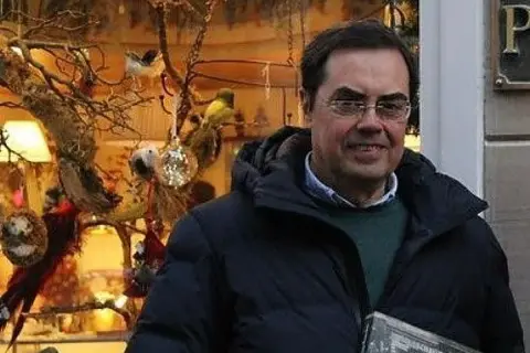 Cayo Pombo Ybarra, sonriendo, con un abrigo oscuro y un suéter verde, frente a un escaparate decorado con pájaros y adornos.
