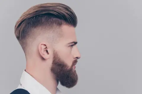 Mężczyzna z zarostem i fryzurą slick back, z wygolonymi bokami i dłuższymi włosami na górze.