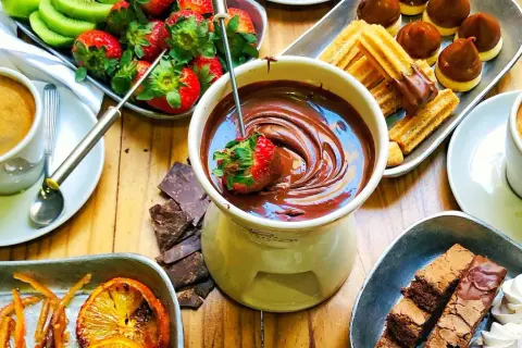Jak przygotować czekolady do fontanny: truskawka zanurzona w płynnej czekoladzie, obok kawa, ciastka, owoce i brownie.