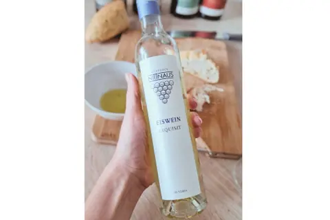 Dłoń trzyma butelkę lodowego wina Eiswein Exquisit z Austrii. Na stole leży chleb i miseczka z oliwą.