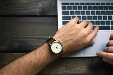 Nadgarstek z zegarkiem i dłonie na klawiaturze laptopa symbolizują nienormowany czas pracy.