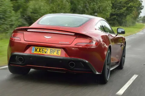 Czerwony Aston Martin Vanquish pędzi po mokrej drodze, otoczony zielenią.