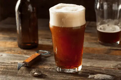 Un bicchiere di birra rossa con schiuma abbondante, accanto a una bottiglia e un apribottiglie. Perfetta per gustare i suoi gradi.