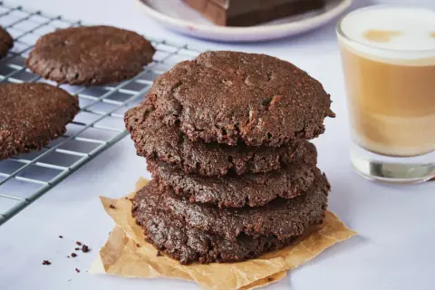 Ein Stapel saftiger brownie cookies, daneben ein Glas Latte Macchiato und ein Teller mit Schokoladenstücken.