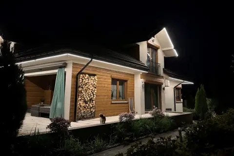 Nowoczesne oświetlenie w podbitce odległości podkreśla architekturę domu. Widać stos drewna, małego psa i kwitnące krzewy.