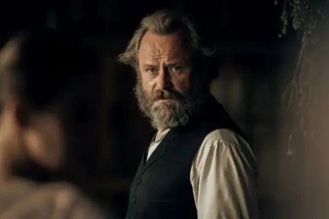 Mężczyzna z filmu Znachor, z siwą brodą i zmierzwionymi włosami, stoi w mrocznym wnętrzu. Gdzie jest młyn z filmu Znachor?