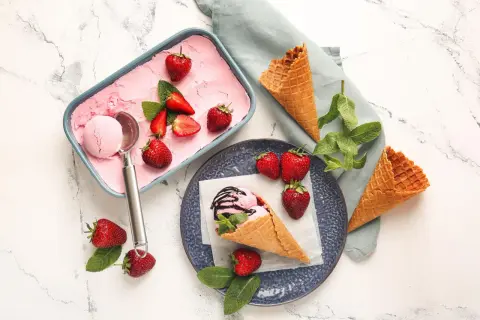 Erdbeer-Eis in einer Schale, garniert mit frischen Erdbeeren und Minze. Daneben leere Waffeln und eine Eiskugel im Hörnchen.