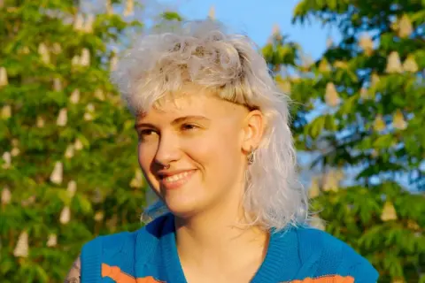 Ragazza con un audace mullet donna lungo, capelli biondi cotonati e rasati ai lati, sorride con gioia sotto un cielo azzurro, circondata da alberi in fiore.