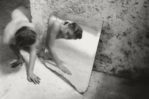Une femme se contorsionne près d'un miroir brisé, son reflet fragmenté. Une œuvre de Francesca Woodman.