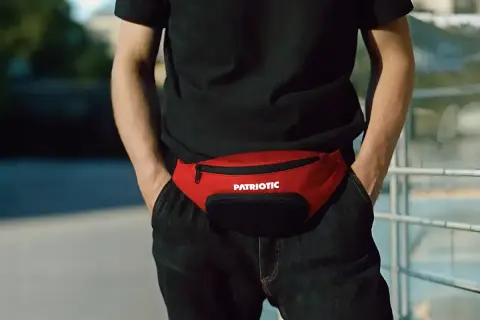 Mężczyzna w czarnej koszulce i jeansach nosi czerwoną nerkę z napisem "PATRIOTIC". Stylowy sposób na to, jak nosić nerkę męską.