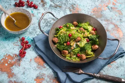 Wirsingsalat mit Fleischbällchen und roten Johannisbeeren in einer Metallschüssel, daneben eine Schüssel mit Dressing.