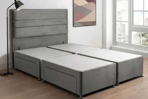 A grey sprung divan base with drawers, paired with a matching headboard, stands in a bright room with a large window.