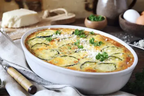 Zapiekanka z cukinii w białym naczyniu, ozdobiona natką pietruszki i parmezanem.