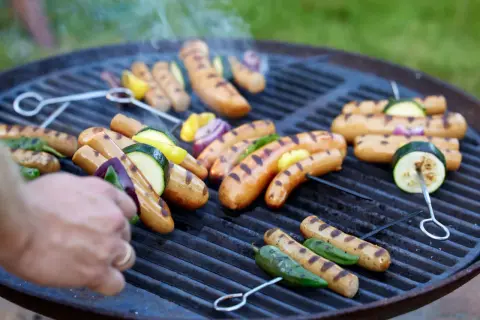 Grilla korv med grönsaker på en kolgrill. Röken stiger från de saftiga korvarna och grönsakerna som får fina grillränder.