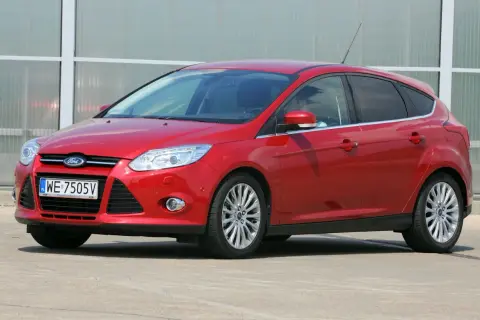 Czerwony Ford Focus z włączonymi światłami mijania, zaparkowany przed nowoczesnym budynkiem.